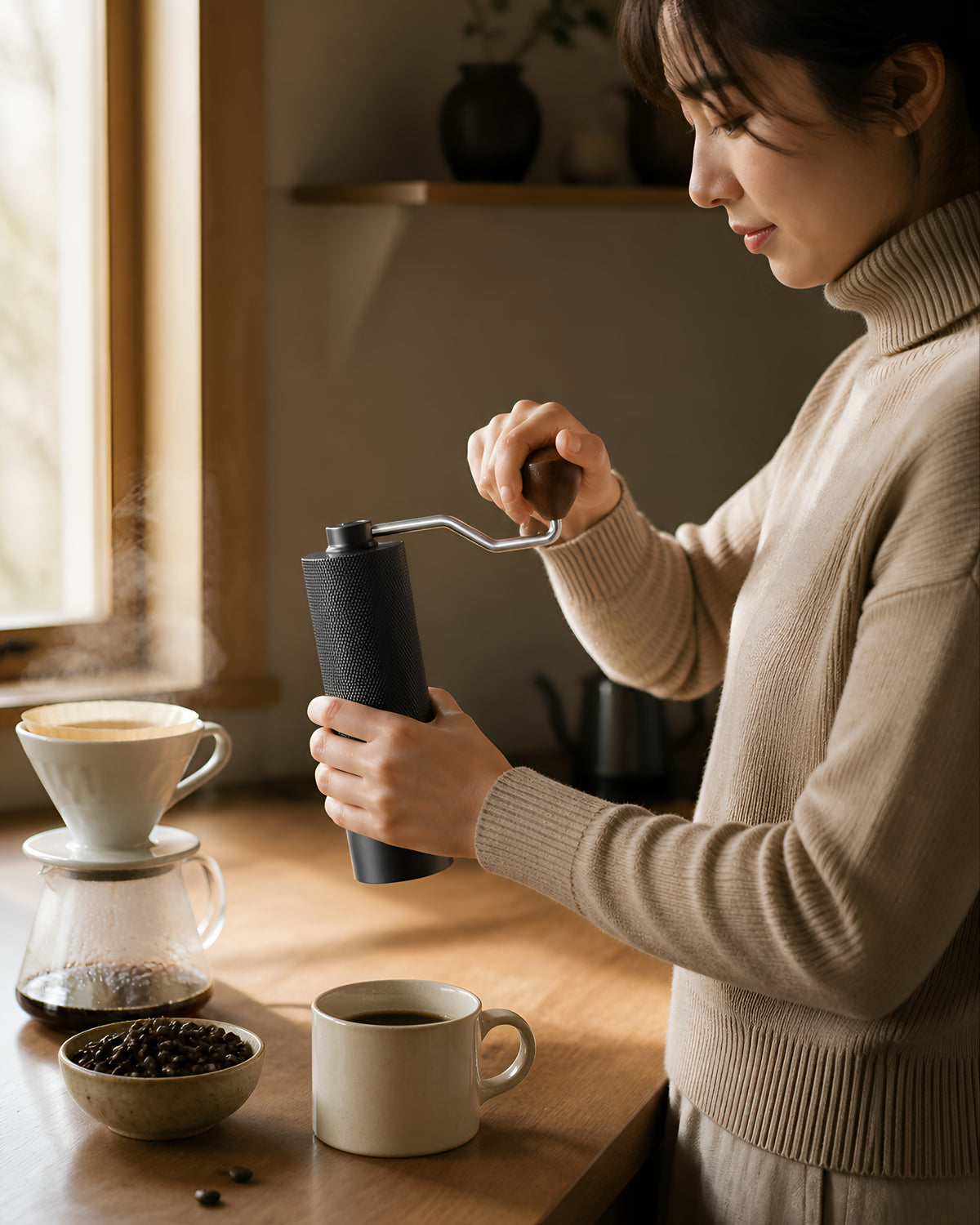 Person using a black manual coffee grinder beside a pour-over brewer in a kitchen